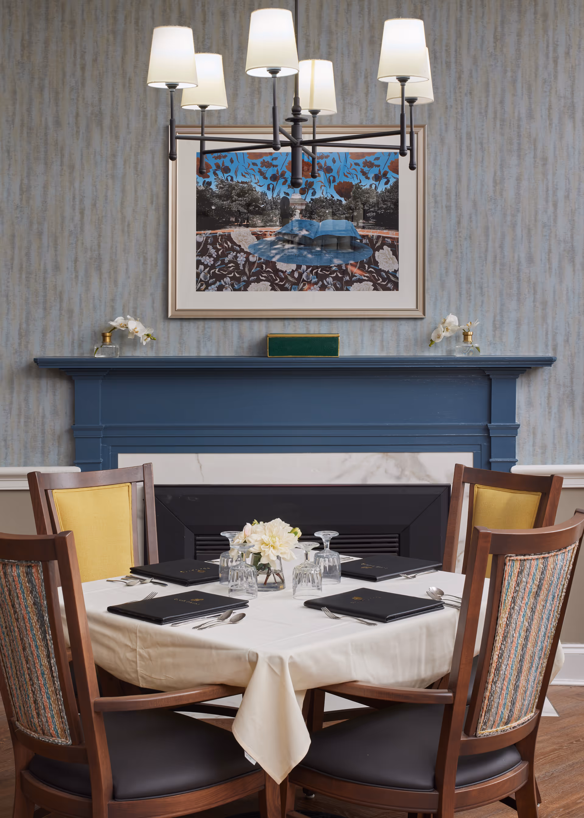 A dining table set for four with menus, glasses, and silverware in a room with a blue fireplace mantel, a floral centerpiece, a modern chandelier with five lamps, and a framed artwork on the wall.