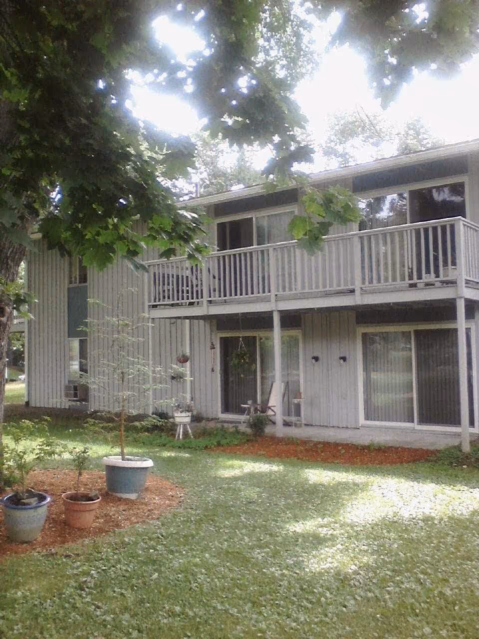 Two-story retirement building with an upper balcony, sliding glass doors, and a grassy yard with potted plants.