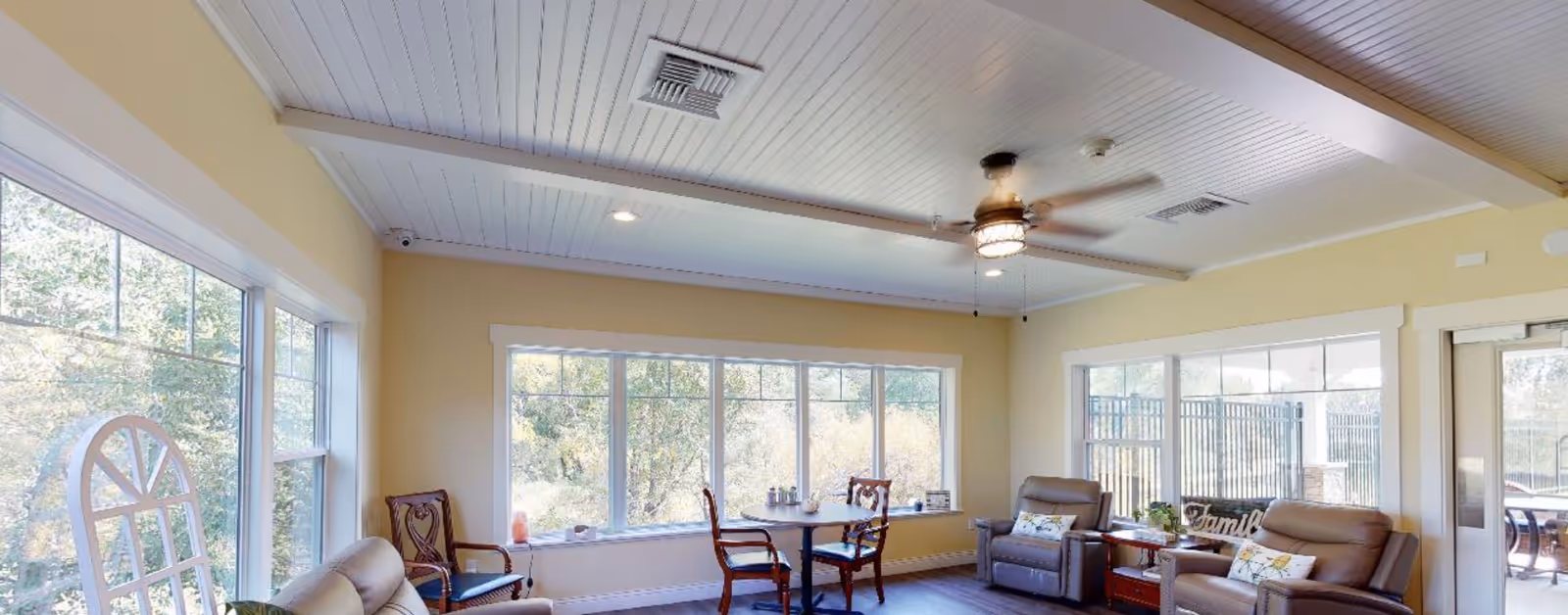 A bright and cozy living room with large windows letting in natural light. The room features beige walls, a white paneled ceiling with exposed beams, a ceiling fan, two leather recliners with decorative pillows, a small round table with two wooden chairs, and a wooden side table with plants and a decorative 'Family' sign. Outside the windows, greenery is visible.