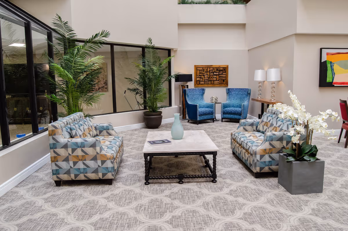 A bright and spacious senior living community lounge area with patterned armchairs, two blue upholstered chairs, a square coffee table with a vase, large potted plants, a side table with two lamps, and modern artwork on the walls.