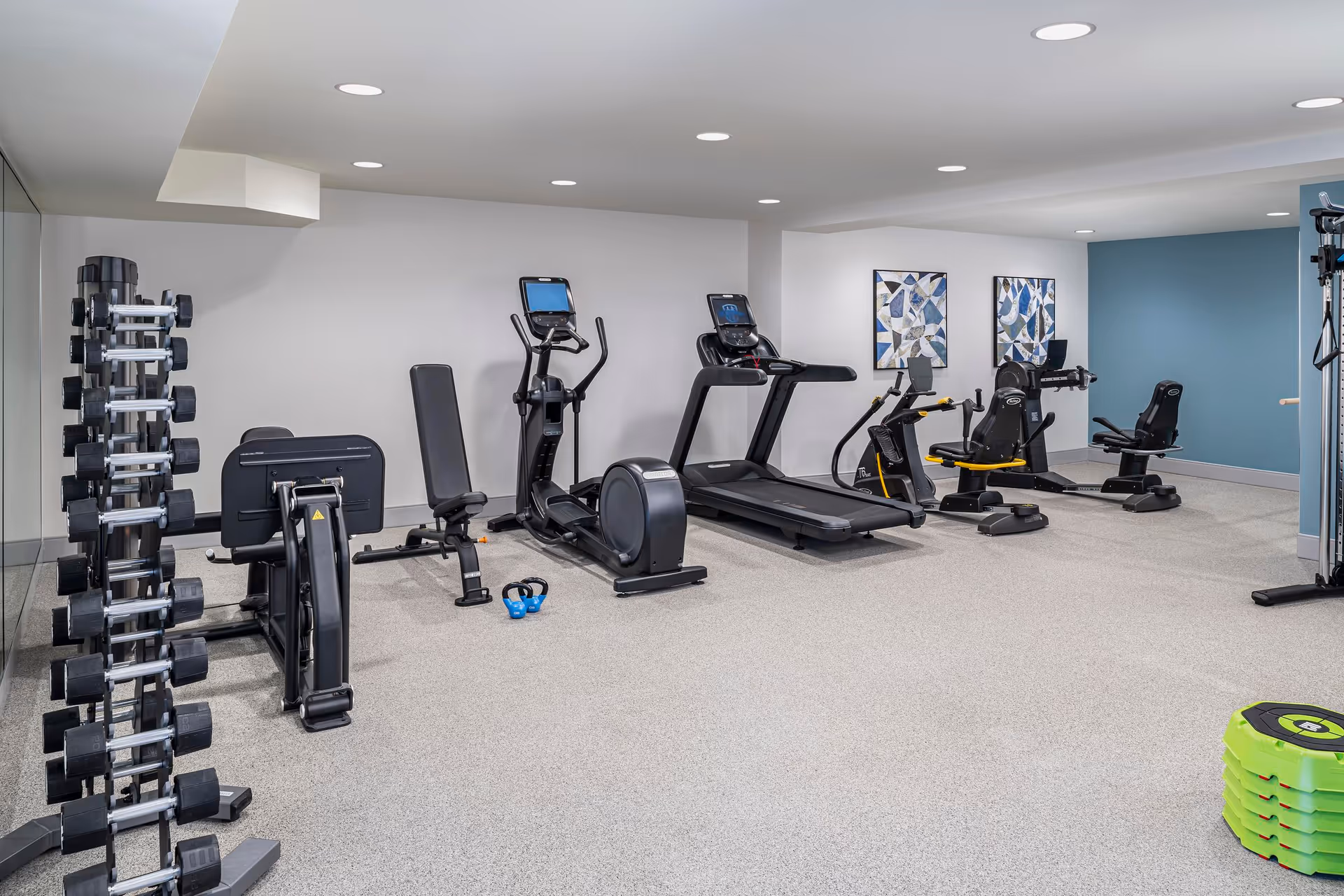 Small fitness room with treadmills, ellipticals, recumbent bikes, and a dumbbell rack.