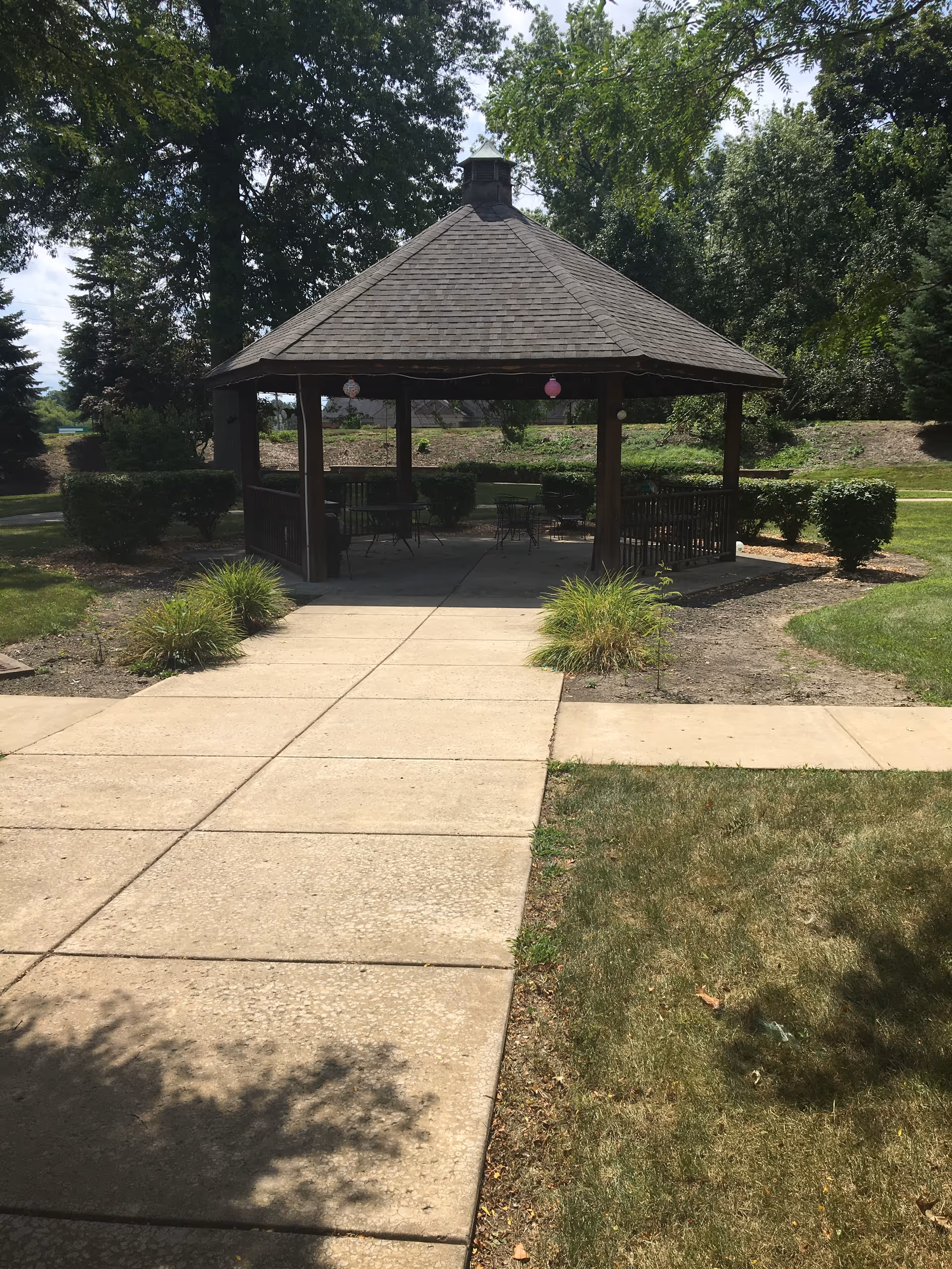 A wooden gazebo with a shingled roof situated at the end of a concrete walkway, surrounded by green grass, bushes, and trees under a partly cloudy sky.
