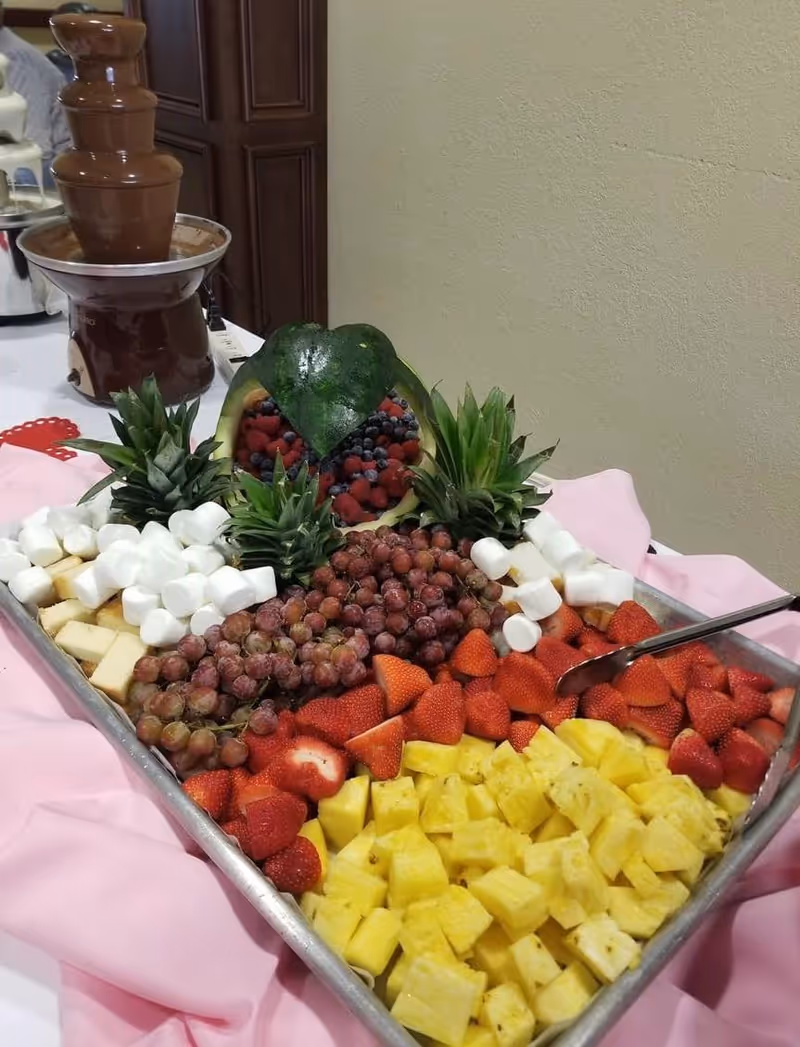 A tray of assorted fresh fruits including pineapple chunks, strawberries, grapes, and marshmallows arranged neatly with a decorative watermelon bowl filled with berries and topped with a carved watermelon leaf. A chocolate fountain is visible in the background.