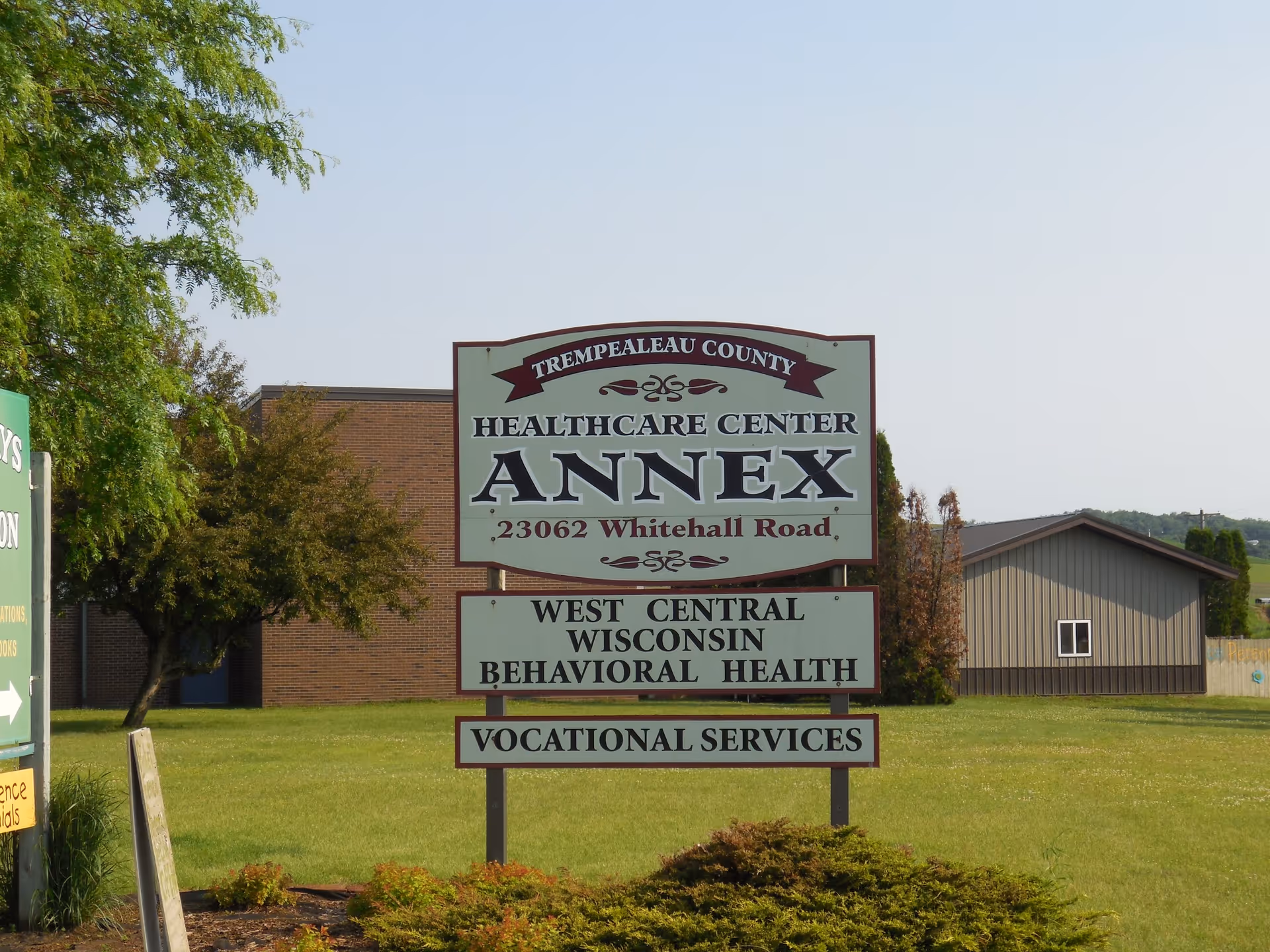 Outdoor view of a sign for Trempealeau County Health Care Center Annex located at 23062 Whitehall Road, with additional signs for West Central Wisconsin Behavioral Health and Vocational Services. The sign is set on a grassy area with trees and buildings in the background under a clear sky.