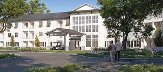 Exterior view of a large, white senior living facility building with multiple windows and a covered entrance. There are trees and landscaped greenery around the building, and two people walking on a pathway in front of it.