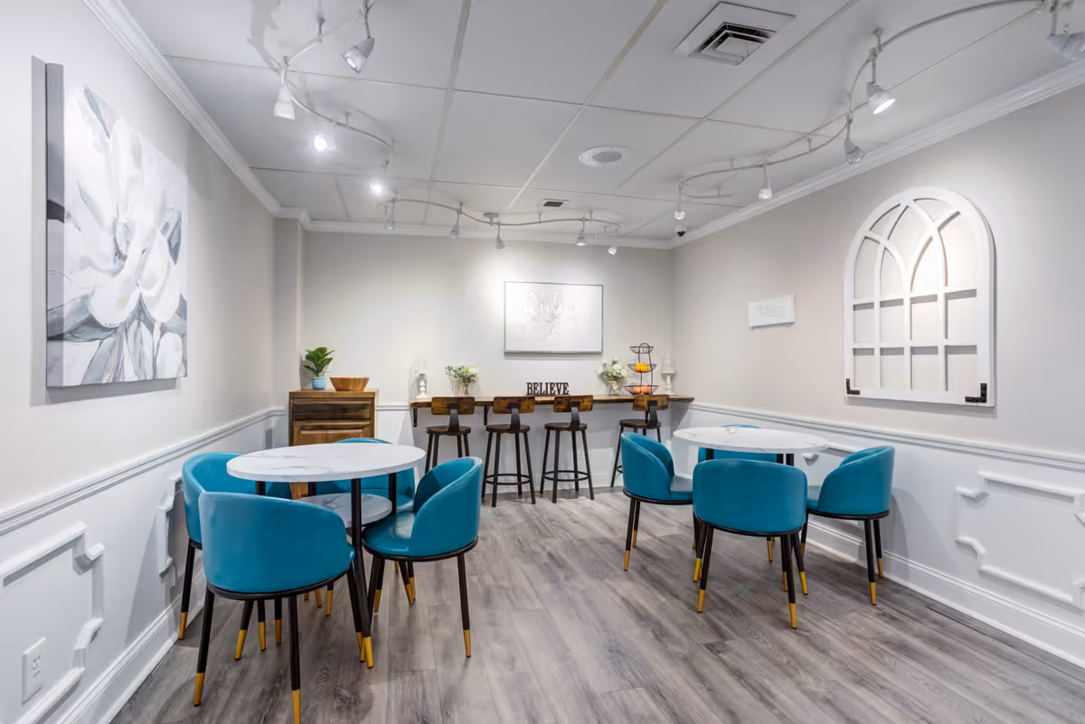 A bright, modern dining area with two round white tables surrounded by teal chairs with black legs and gold tips. The room has light gray walls with white wainscoting and wood-look flooring. On the back wall, there is a wooden counter with four high stools, decorative flowers, a tiered fruit basket, and a sign that says 'BELIEVE'. The walls are decorated with light-colored floral artwork and a white arched window frame. Ceiling track lighting illuminates the space.