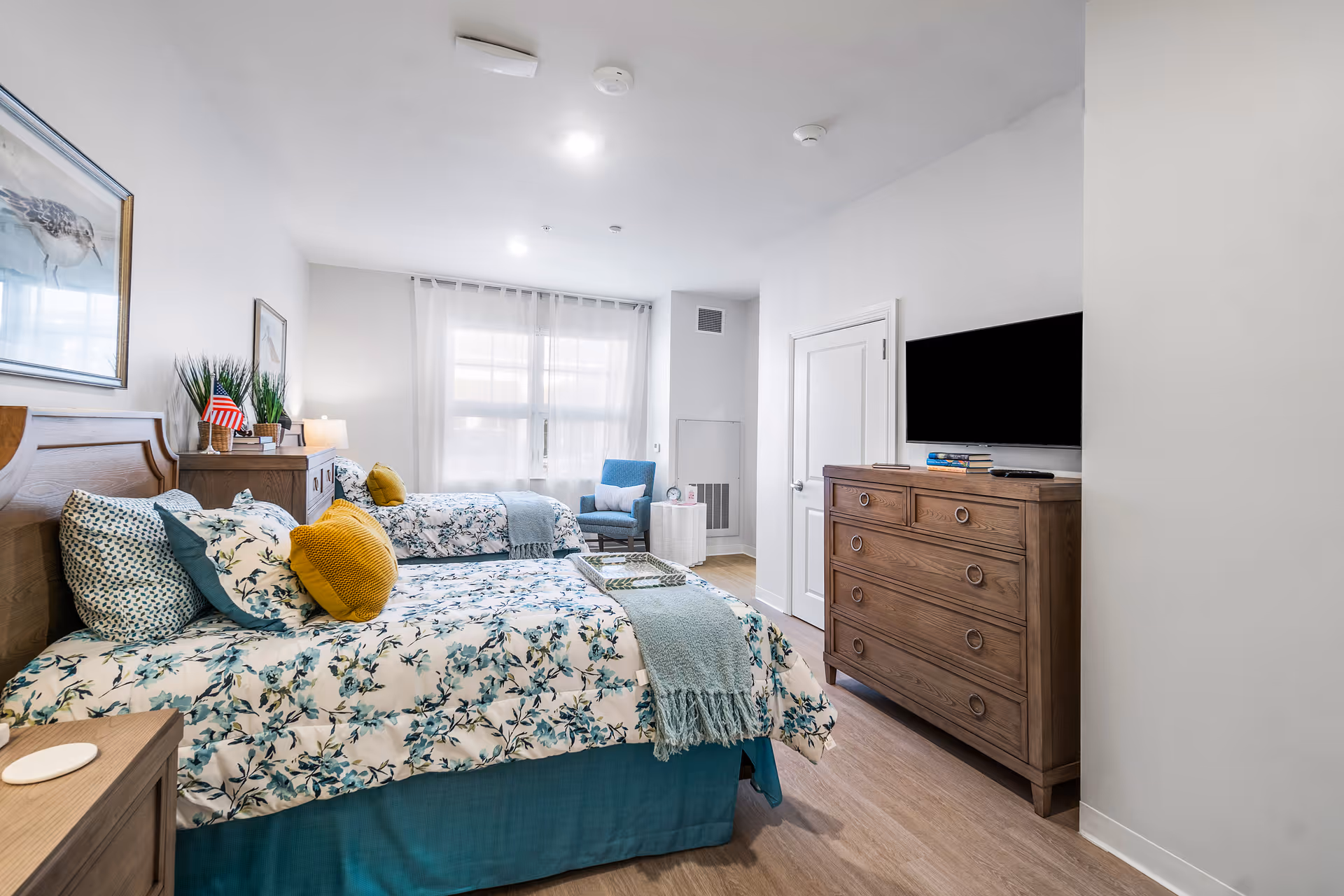 A bright and clean bedroom with two twin beds featuring floral bedding and yellow accent pillows. There is a wooden dresser with a flat-screen TV mounted above it, a blue armchair near a window with sheer white curtains, and framed artwork on the walls. The room has light wood flooring and white walls.