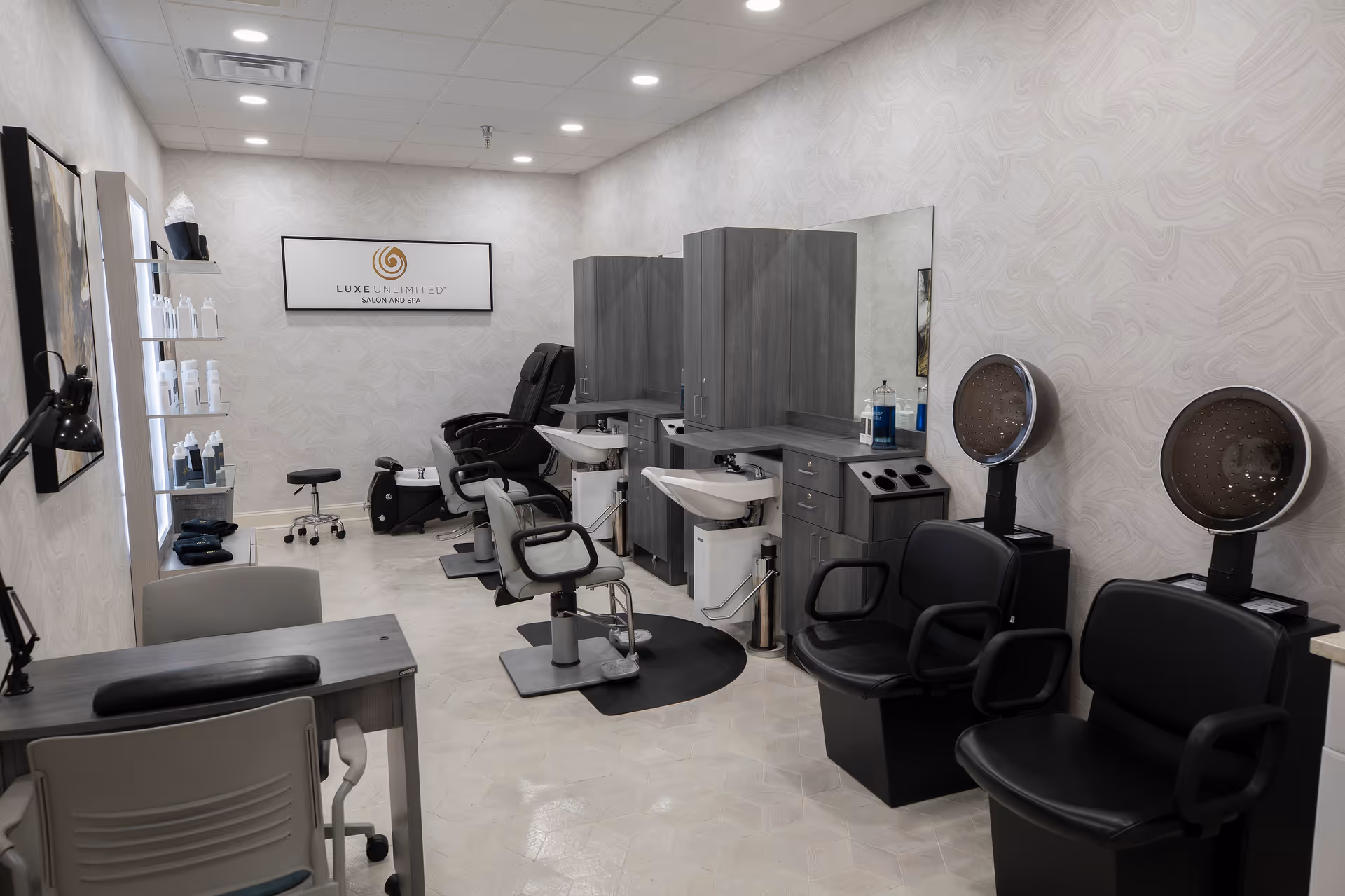 Interior view of a salon and spa room with two hair washing stations, two salon chairs in front of mirrors, two hair dryers with chairs, a manicure table with chairs, and shelves with hair care products. The room has light-colored walls and ceiling with recessed lighting.