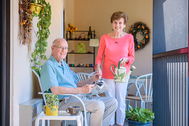 Two older adults on a sunlit balcony holding a watering can and a newspaper surrounded by potted plants and patio furniture.
