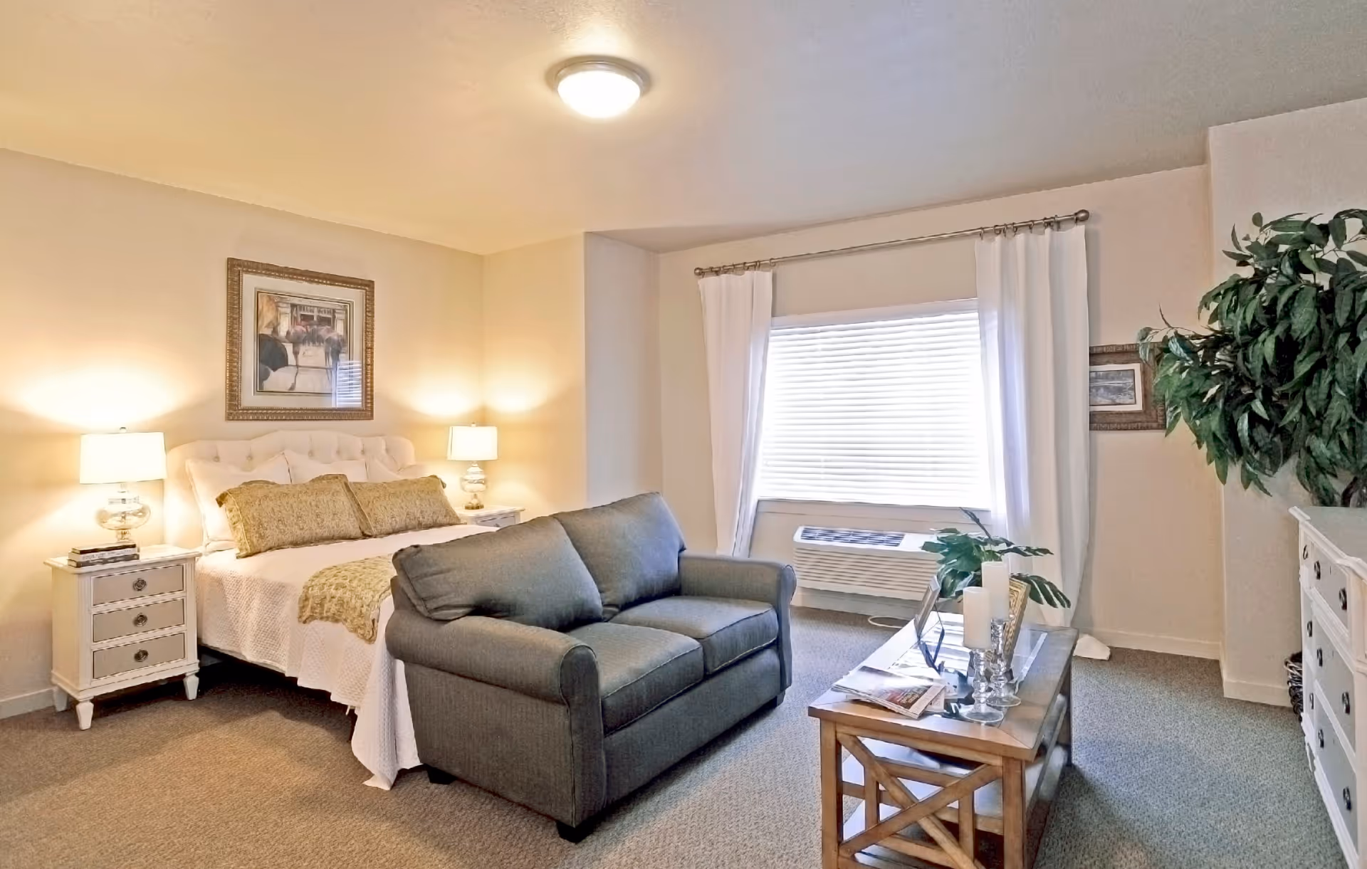 A well-lit senior living bedroom featuring a large bed with white bedding and decorative pillows, flanked by two nightstands with lamps. In front of the bed is a gray loveseat, and a wooden coffee table with decorative items. A large window with white curtains and blinds is on the right wall, with an air conditioning unit below it. A tall green plant and a white dresser are also visible in the room.