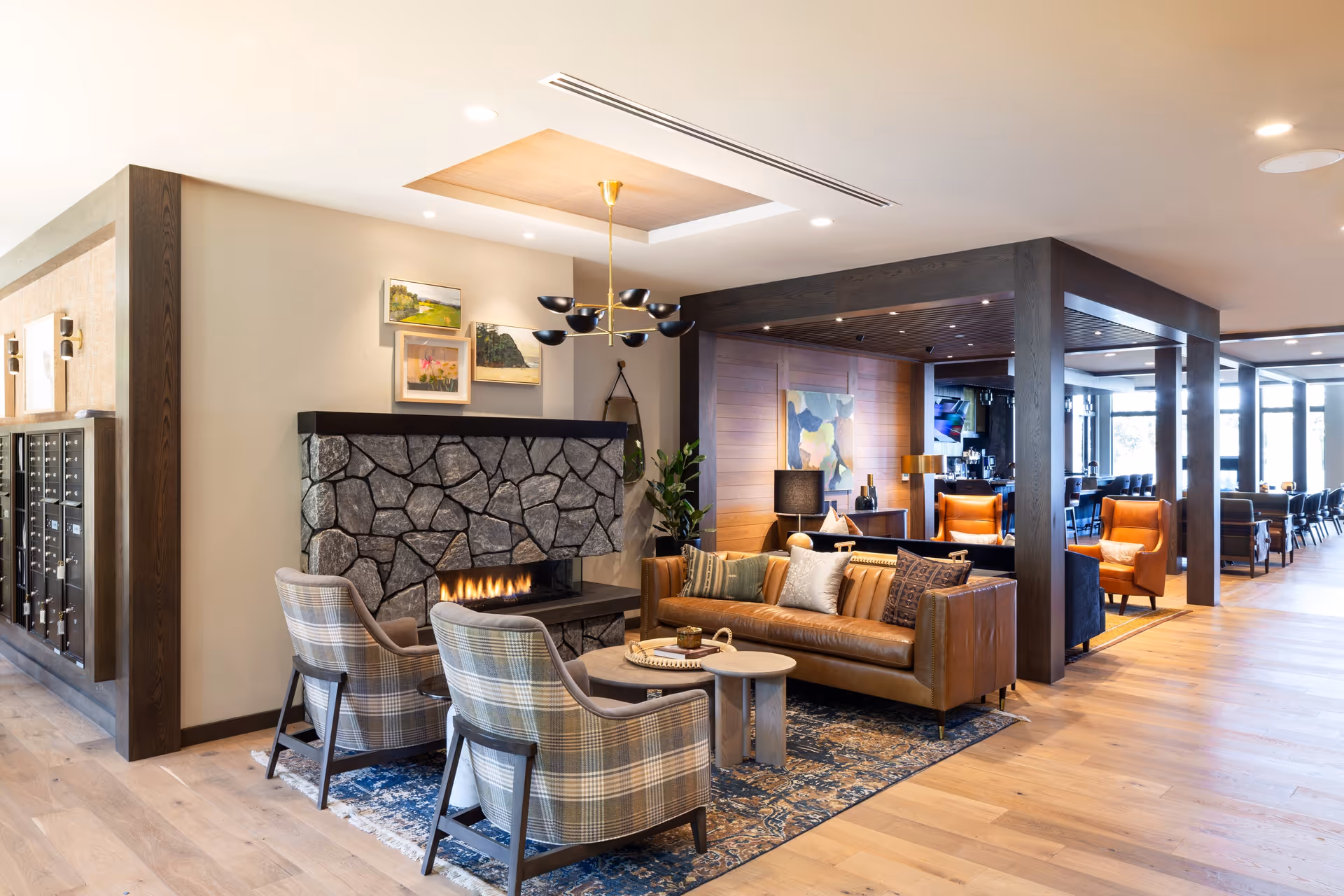 Cozy communal lounge with a stone fireplace, leather sofa, plaid armchairs, and modern lighting.