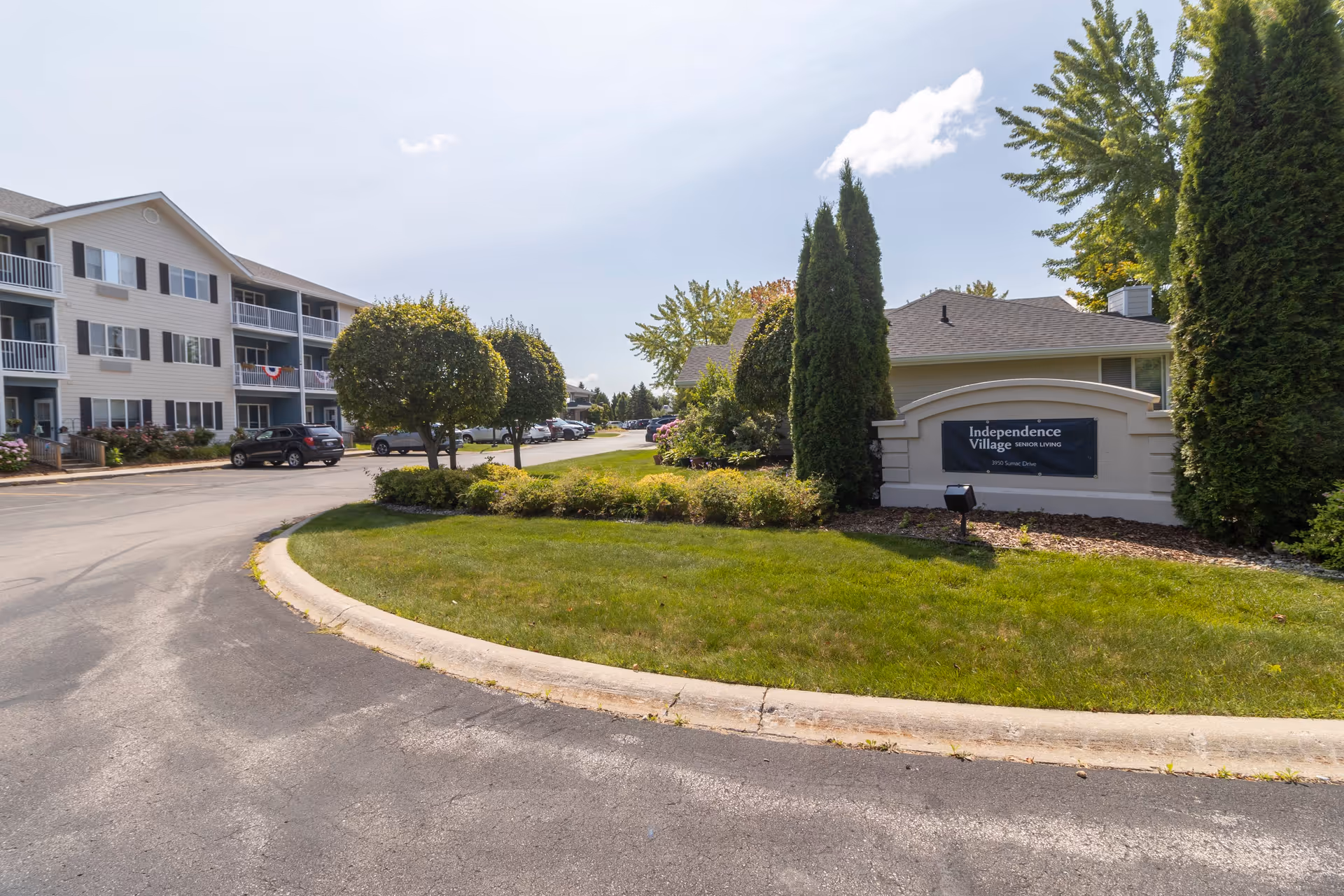 Exterior view of Independence Village of Traverse City senior living facility showing a curved driveway, manicured lawn, trimmed bushes, and a sign with the facility's name. A multi-story residential building with balconies and parked cars is visible on the left side under a partly cloudy sky.