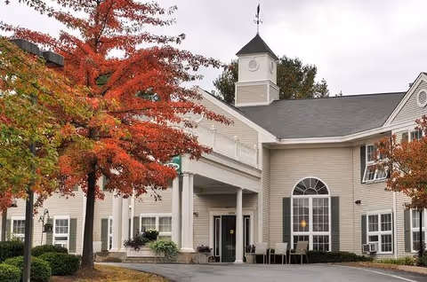 Front entrance of a beige senior living building with white columns, a cupola, and red autumn trees.