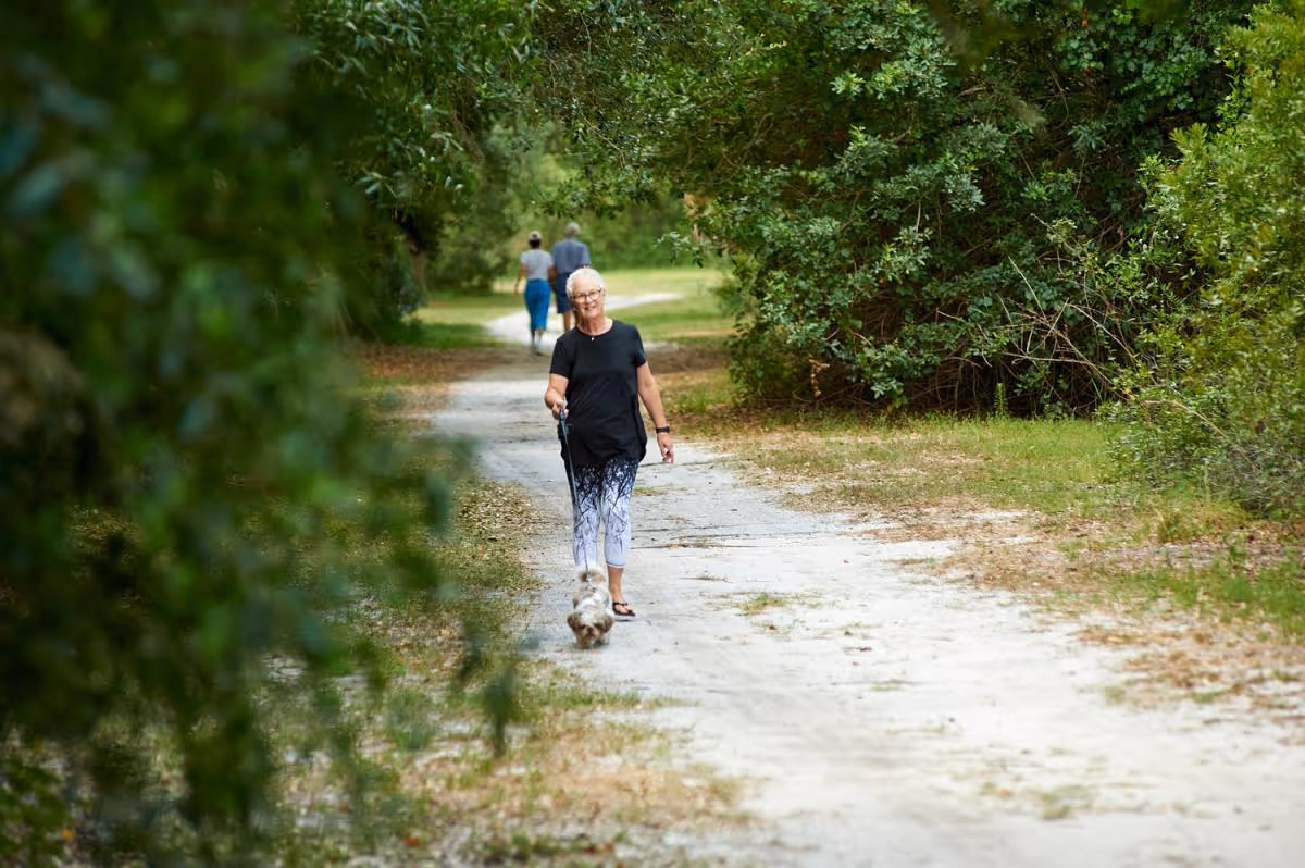 An elderly woman walking a small dog on a leash along a dirt path surrounded by green trees and bushes. Two other people are walking further ahead on the same path.