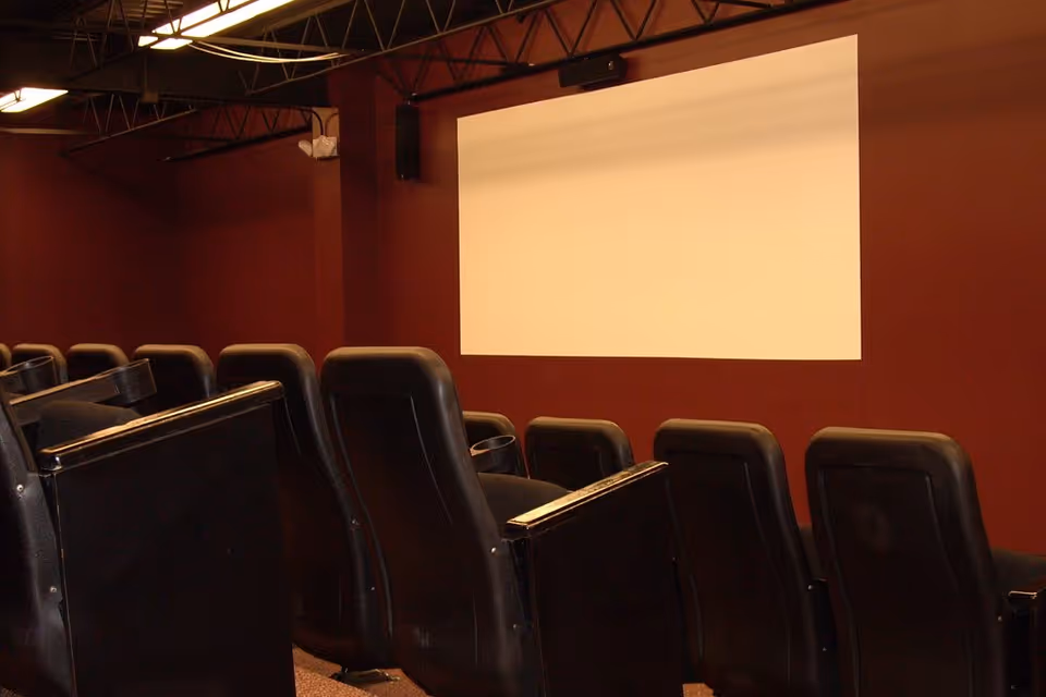 Interior view of a small theater room with rows of black cushioned seats facing a blank white projection screen on a maroon wall.