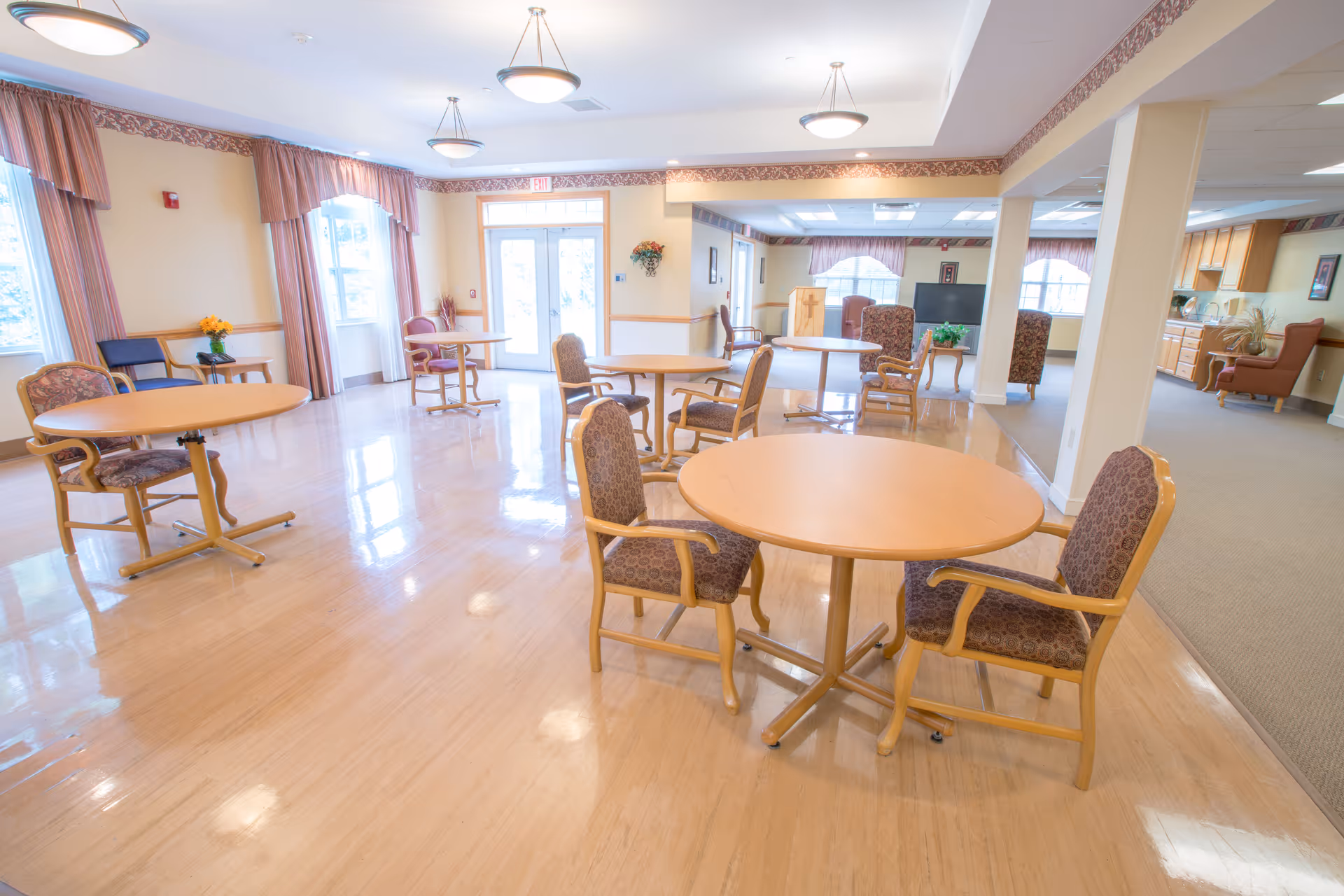 A bright and spacious common area in McNaughten Pointe featuring several round wooden tables with cushioned chairs arranged around them. The room has large windows with pink curtains allowing natural light to fill the space. In the background, there is a seating area with armchairs and a television, as well as a kitchenette area with cabinets and countertops. The floor is polished wood, and the walls are painted in light colors with decorative trim near the ceiling.
