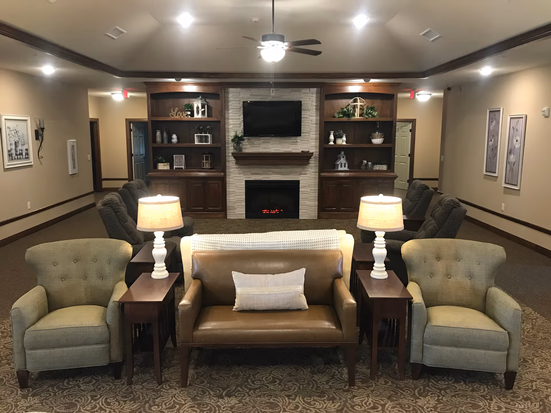 A cozy living room area with a brown leather loveseat, two green upholstered armchairs, and two side tables each holding a lamp. In the background, there are two wooden built-in shelves flanking a white stone fireplace with a flat-screen TV mounted above it. The room has beige walls, carpeted floor, and ceiling lights with a ceiling fan in the center.