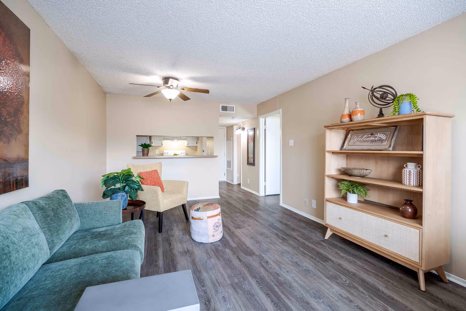 A cozy living room with a green sofa, beige armchair, small round ottoman, and a wooden bookshelf decorated with plants, vases, and a 'Welcome Home' sign. The room has light beige walls, wood flooring, a ceiling fan with light, and a pass-through window to the kitchen.