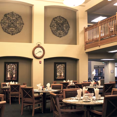 Interior view of a dining room in a senior living facility with multiple round tables set with white napkins, cups, and utensils. The room features high ceilings with decorative wall art, a clock, and a wooden balcony railing on the upper level.