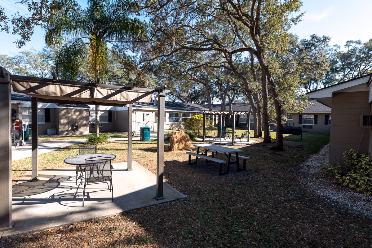 Outdoor courtyard area at The Bristol at Tampa Rehabilitation & Nursing Center featuring a concrete patio with a round metal table and chairs under a pergola, a picnic table on the grass, trees providing shade, and single-story buildings surrounding the space.