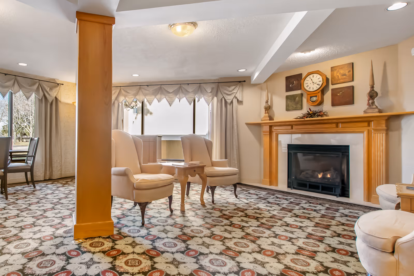 A cozy living room area with two beige armchairs and a small wooden table between them, a fireplace with a wooden mantel decorated with a clock and artwork above it, patterned carpet flooring, and large windows with curtains letting in natural light.