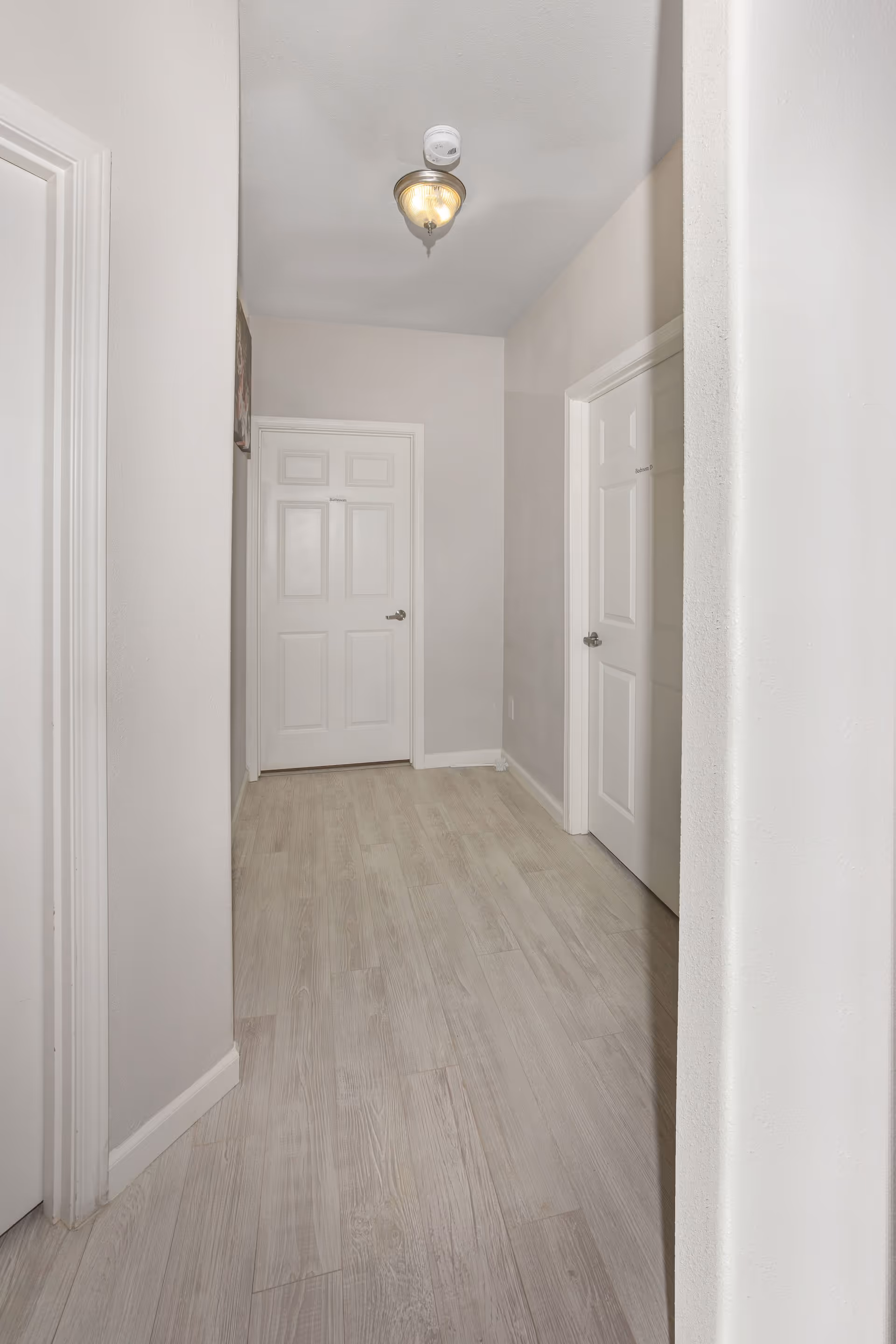 A hallway with light-colored wooden flooring and white walls. There are three white doors visible, one at the end labeled 'Bathroom' and two on the sides, one labeled 'Room 12'. A ceiling light fixture is mounted in the center of the ceiling.
