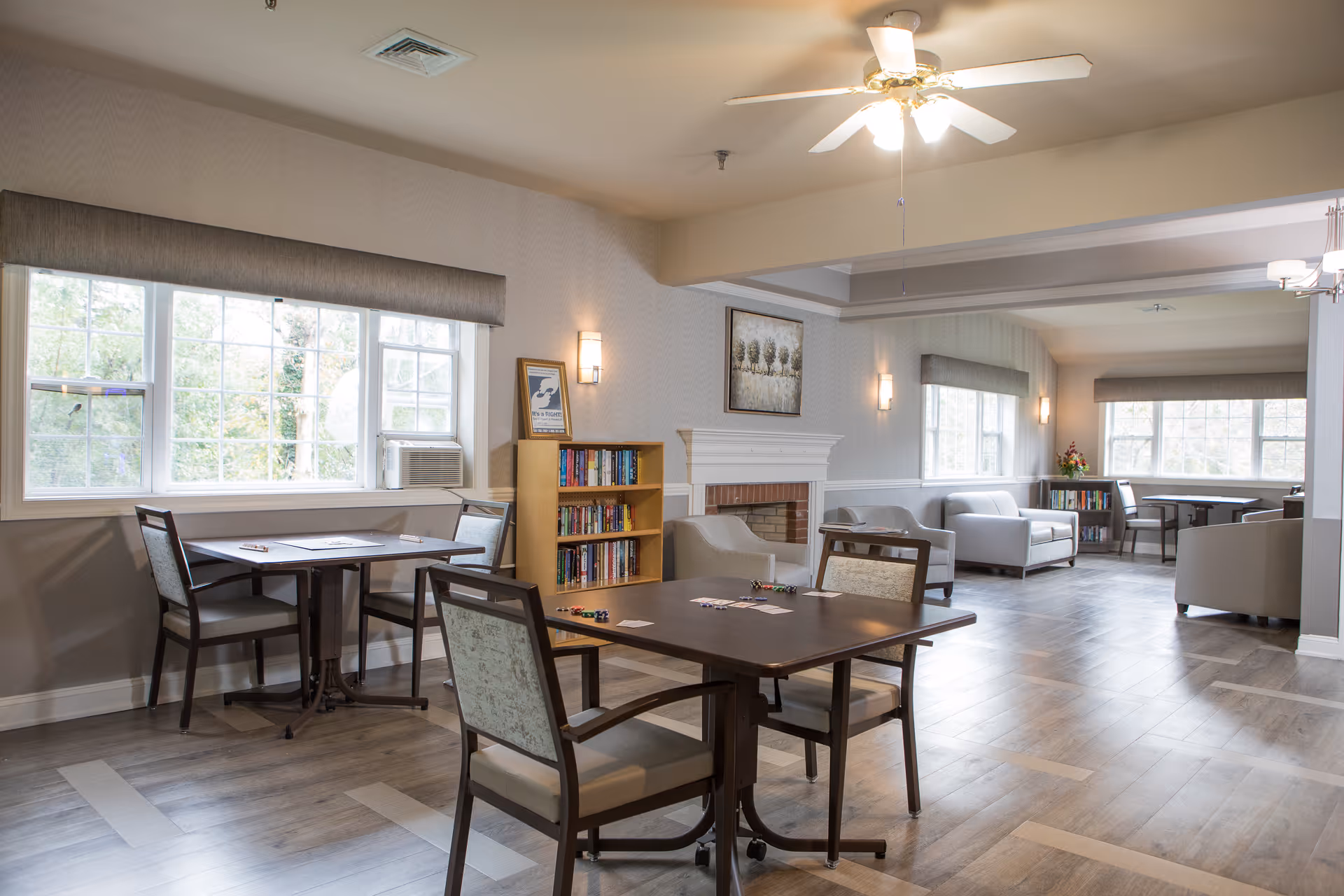 A bright and spacious common area in a senior living facility with multiple seating arrangements including tables with chairs and armchairs. The room features large windows with gray valances, a ceiling fan with lights, a fireplace, bookshelves filled with books, and soft wall lighting. The flooring is wood-style, and the overall decor is neutral and inviting.