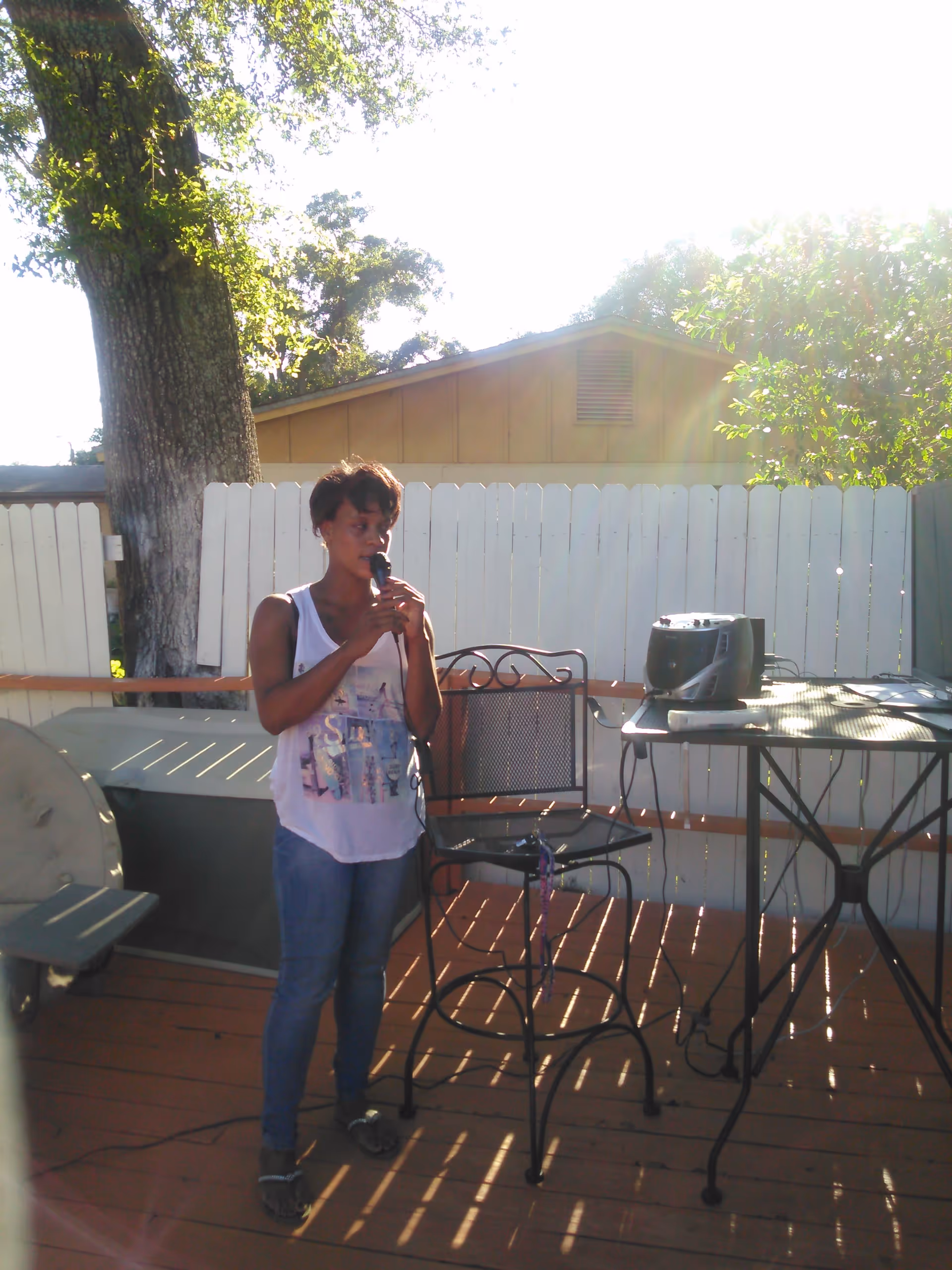 A woman standing on a wooden deck outdoors, holding a microphone and singing or speaking. There is a metal chair and a table with a stereo system and a remote control on it. A white fence and trees are visible in the background with sunlight shining through.
