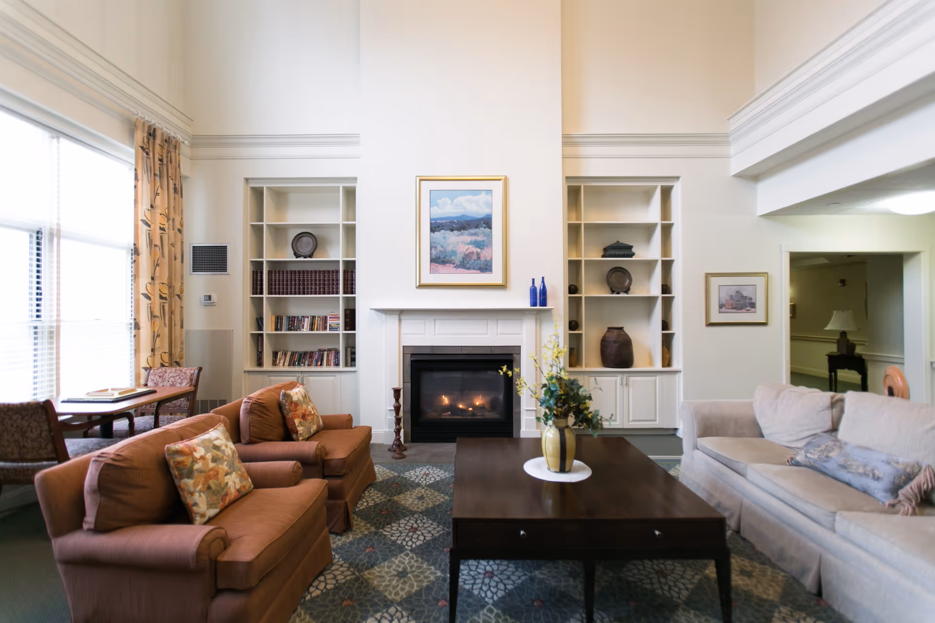 A cozy living room with a fireplace in the center, flanked by built-in white shelves holding books and decorative items. There are two brown armchairs with floral cushions on the left, a beige sofa with a blue pillow on the right, and a dark wooden coffee table with a vase of flowers in the middle. Large windows with patterned curtains allow natural light to fill the room.