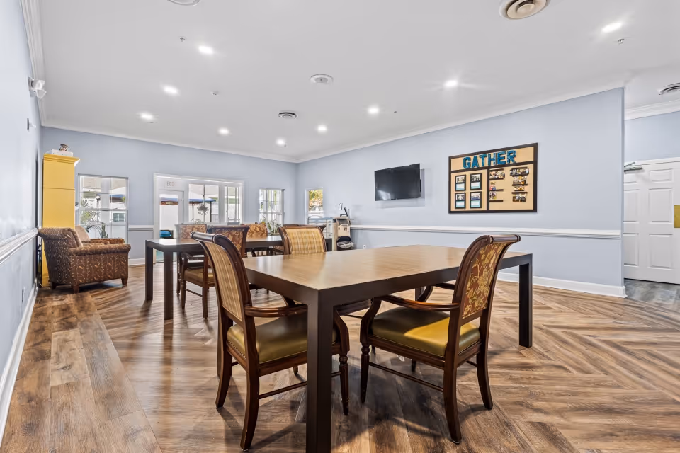 A bright and spacious common area in a senior living facility with wooden flooring, light blue walls, and recessed ceiling lights. The room features a large wooden table surrounded by chairs with patterned upholstery, a wall-mounted television, a bulletin board with the word 'GATHER' and photos, and comfortable armchairs near windows letting in natural light.