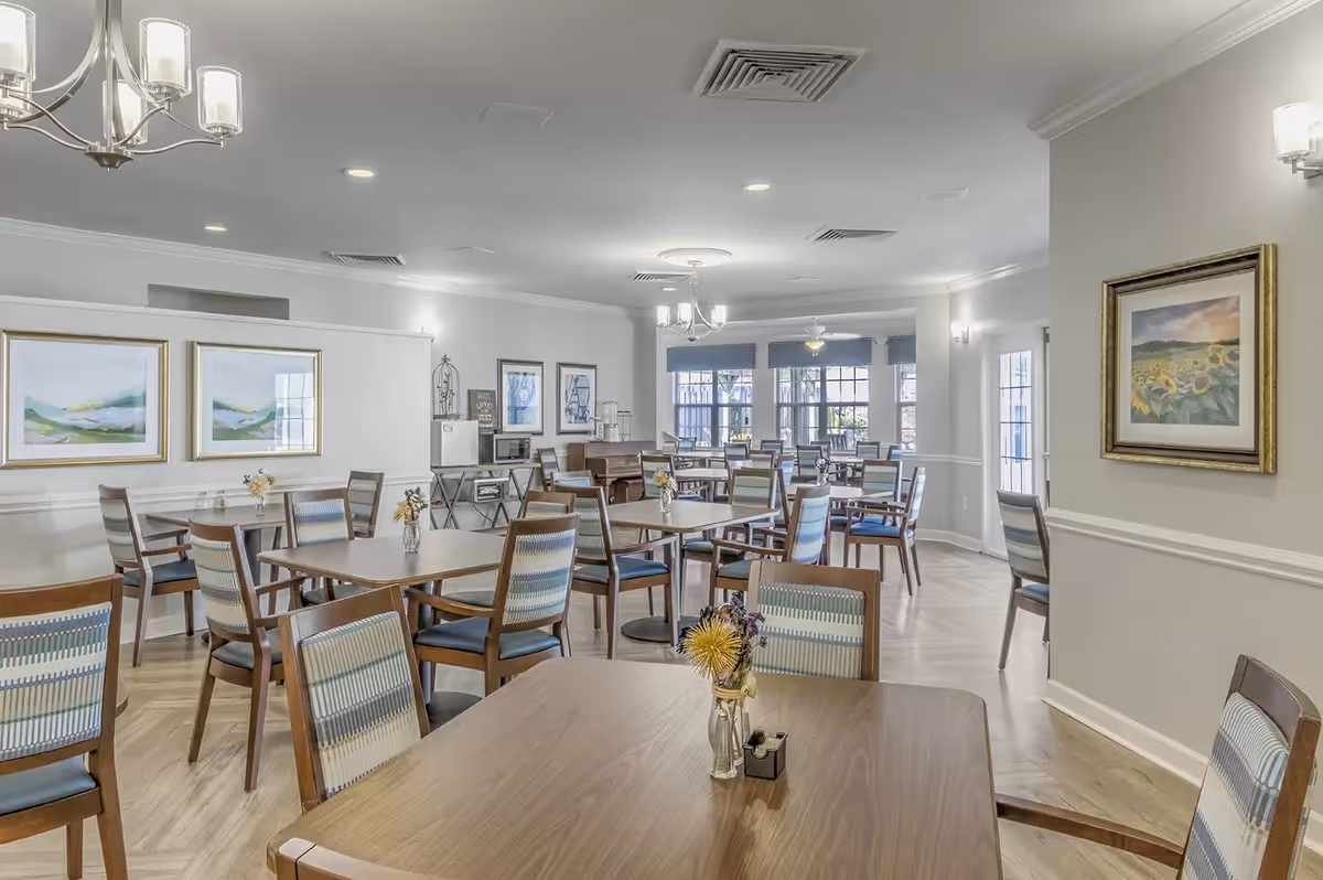 A bright and spacious dining room with multiple wooden tables and chairs arranged neatly. The room features light-colored walls adorned with framed artwork, large windows allowing natural light to fill the space, and modern ceiling lights. Small flower arrangements are placed on the tables, creating a welcoming atmosphere.
