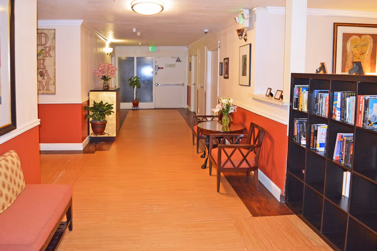 A well-lit hallway in a senior living facility with wooden flooring and red and white walls. There are chairs and small tables with flower vases along the right side, a bookshelf filled with books, and potted plants near the far end. The hallway leads to a set of double doors with an exit sign above.