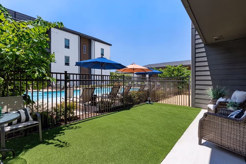 Patio with artificial grass and wicker seating overlooking a fenced pool area with lounge chairs and colorful umbrellas.