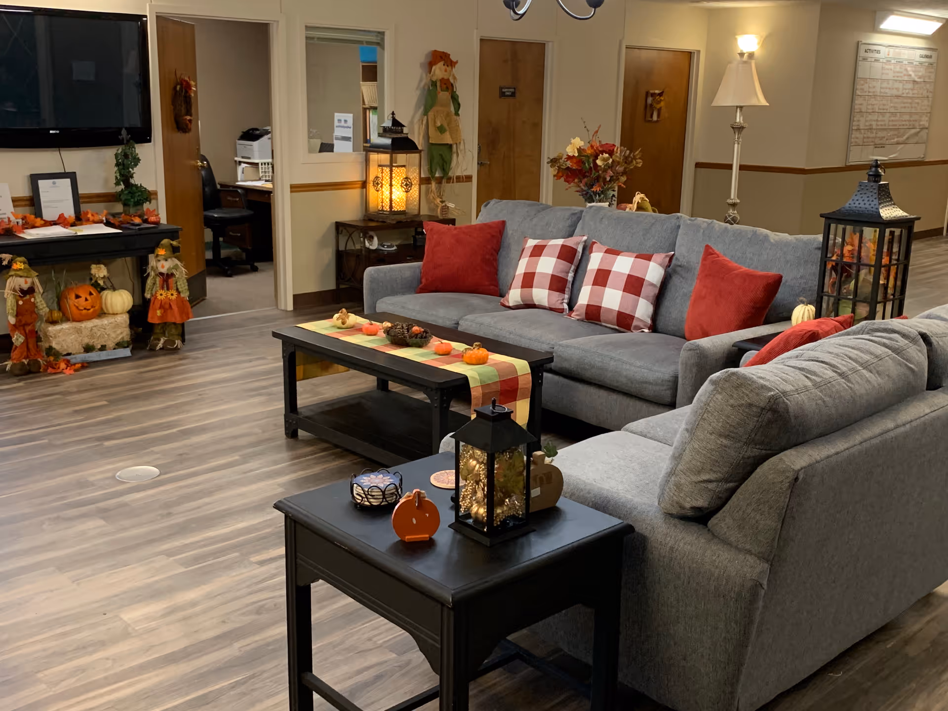 Cozy senior living lounge with gray sofas, red and plaid pillows, coffee tables and fall decorations.