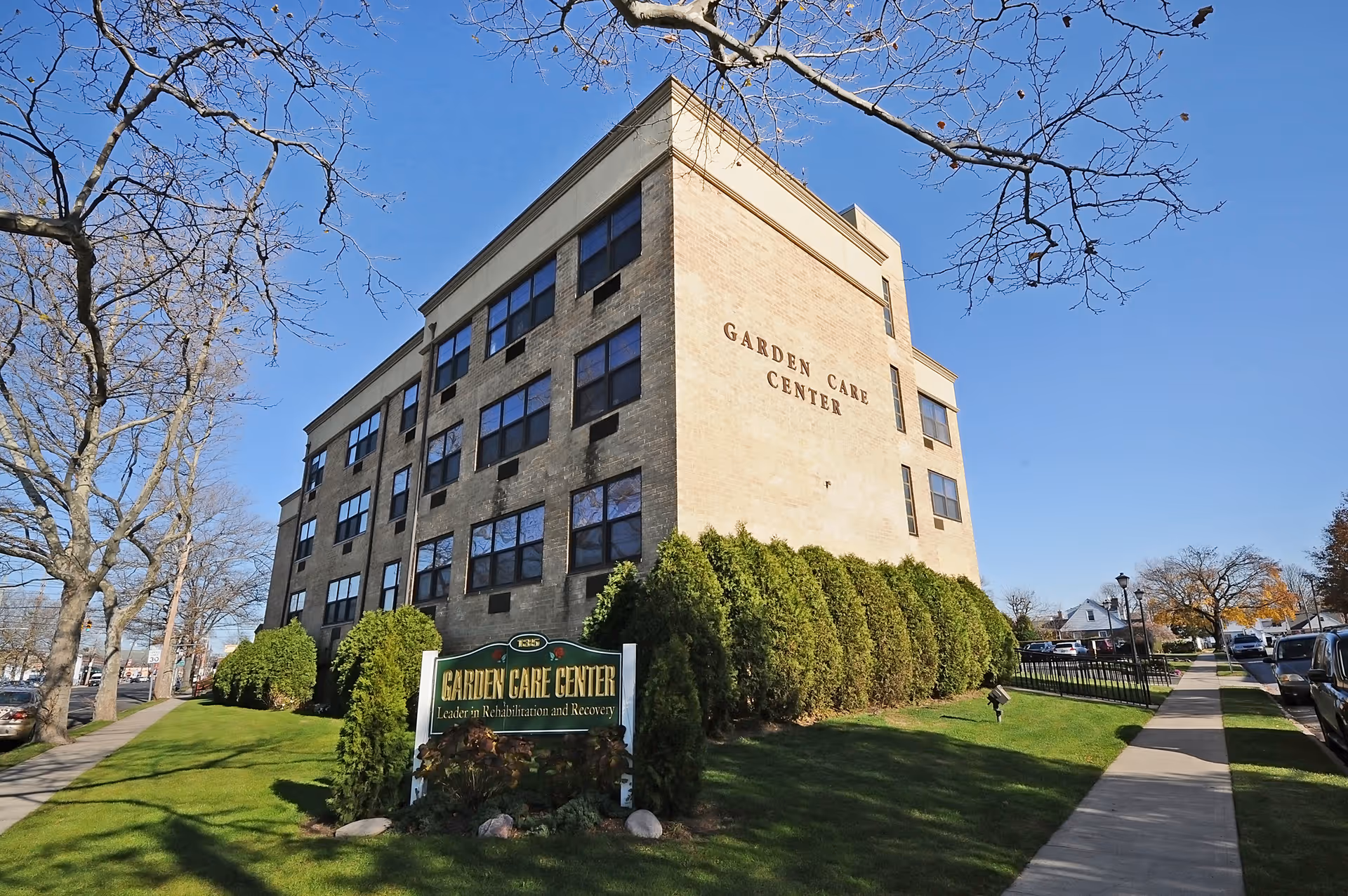 Four-story brick building labeled 'Garden Care Center' with a front lawn, sign, and sidewalk under a clear blue sky.