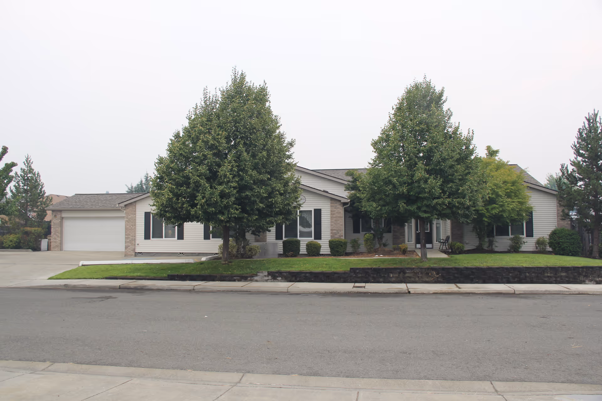 Front exterior view of a single-story residential building with light-colored siding and a two-car garage. The building is partially obscured by two large leafy trees and some shrubs in a well-maintained lawn area. The sky is overcast.
