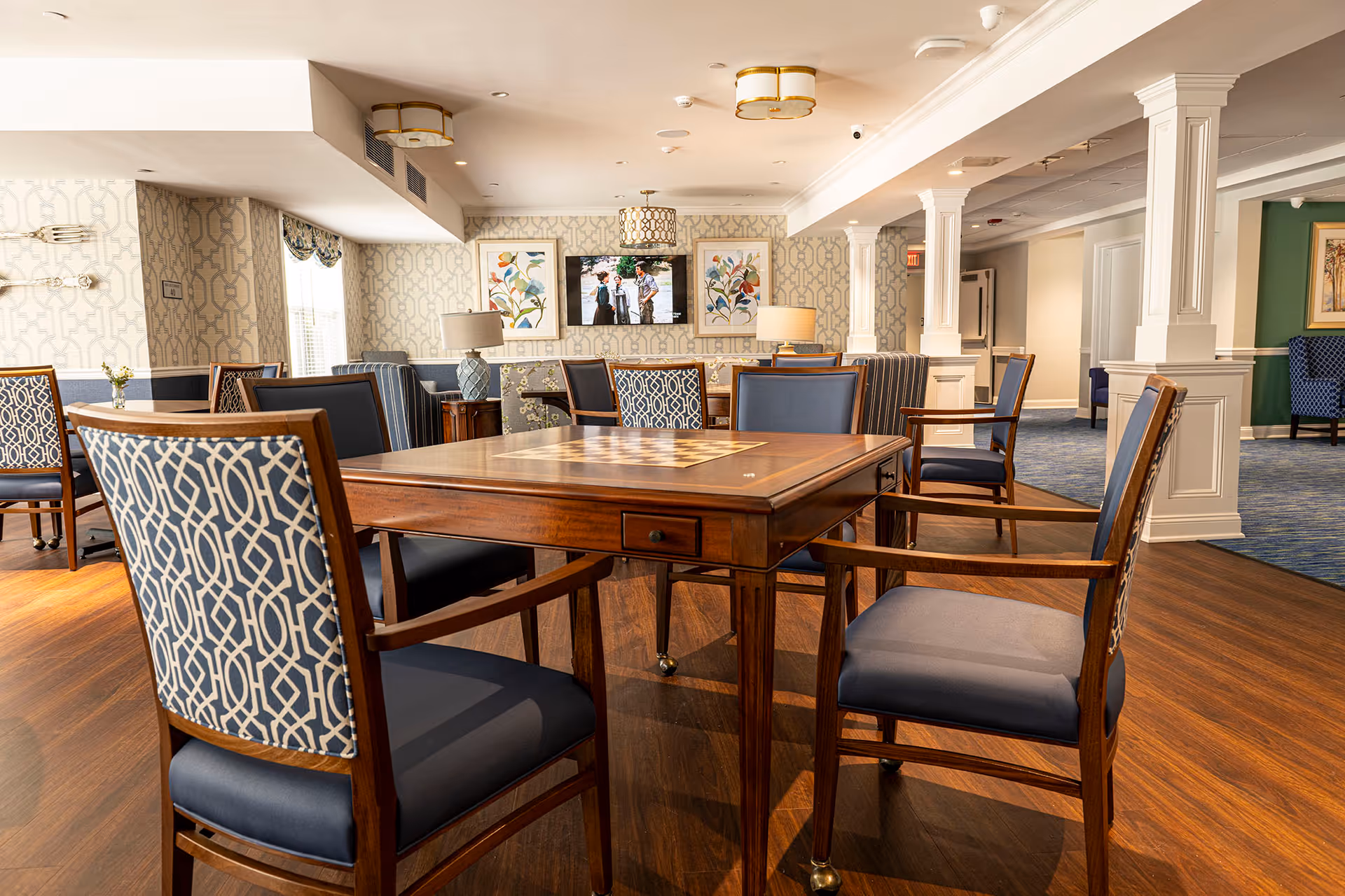 A well-lit common area with a wooden table and several cushioned chairs featuring blue and patterned upholstery. The room has wood flooring, patterned wallpaper, decorative lamps, framed floral artwork, and a television mounted on the wall. White columns and a blue carpeted hallway are visible in the background.