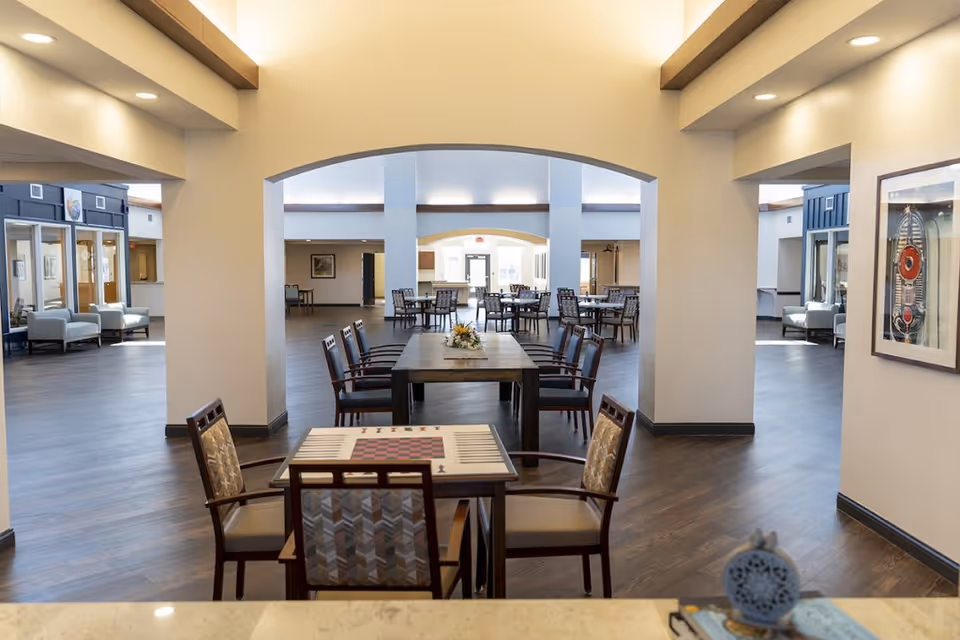 Spacious senior living dining and common area with multiple tables and chairs beneath arched openings and warm wood flooring.