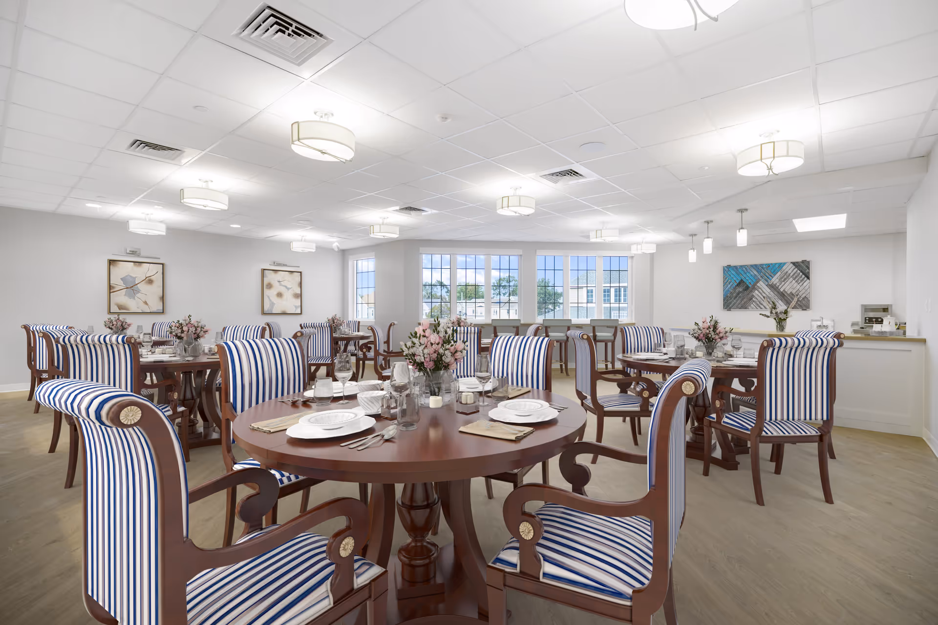 Bright dining room with multiple round wooden tables set with plates, glasses, and cutlery. The chairs have wooden frames with blue and white striped upholstery. There are flower arrangements on the tables, large windows letting in natural light, and modern ceiling lights. Artwork is displayed on the walls and a counter area is visible in the background.