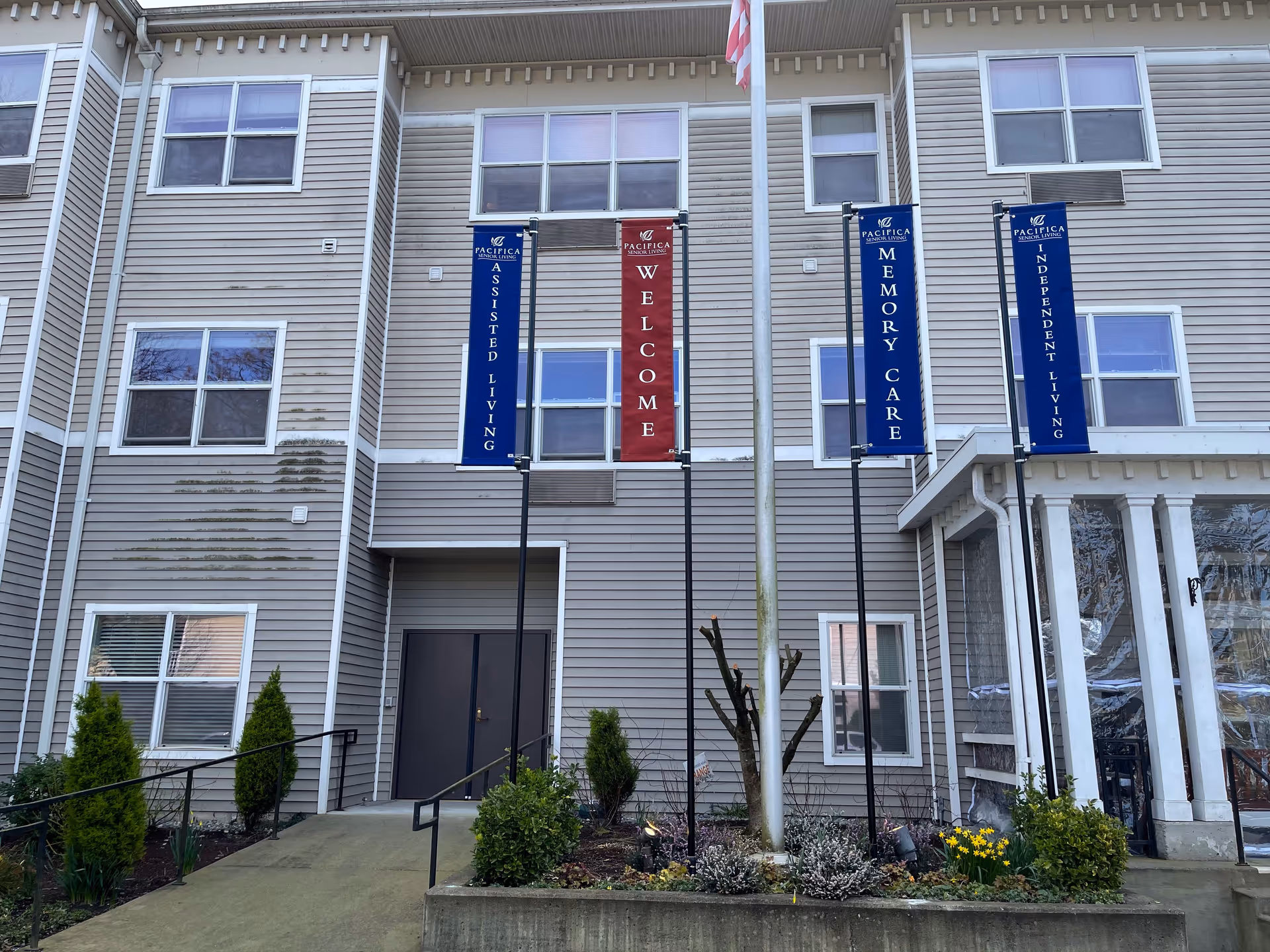 Exterior view of a senior living facility building with beige siding and multiple windows. There are four vertical banners displayed on poles in front of the building. The banners read, from left to right: 'Assisted Living', 'Welcome', 'Memory Care', and 'Independent Living'. There is a small garden with shrubs and flowers at the base of the poles, and a concrete walkway leading to a double door entrance.