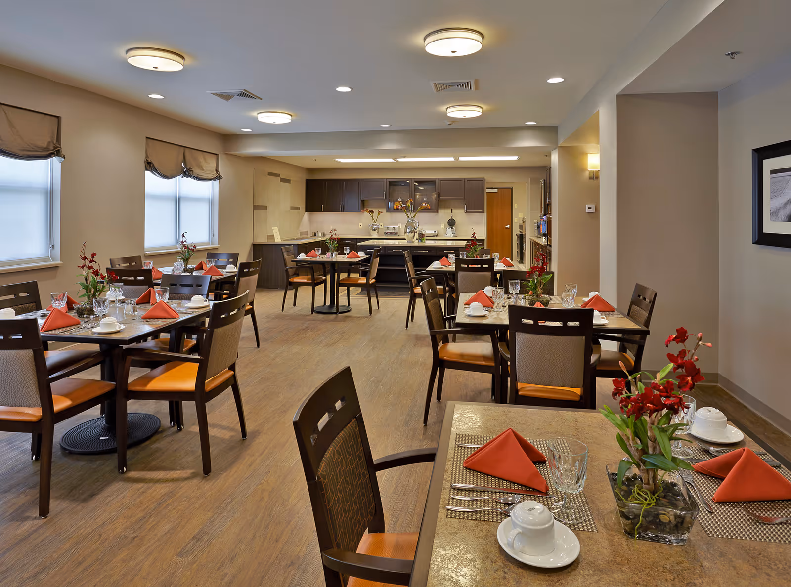 A dining room in a senior living facility with several tables set with red folded napkins, white cups and saucers, glassware, and small floral centerpieces. The room has wooden chairs with cushioned seats, large windows with beige roman shades, and a kitchen area with dark cabinets in the background.