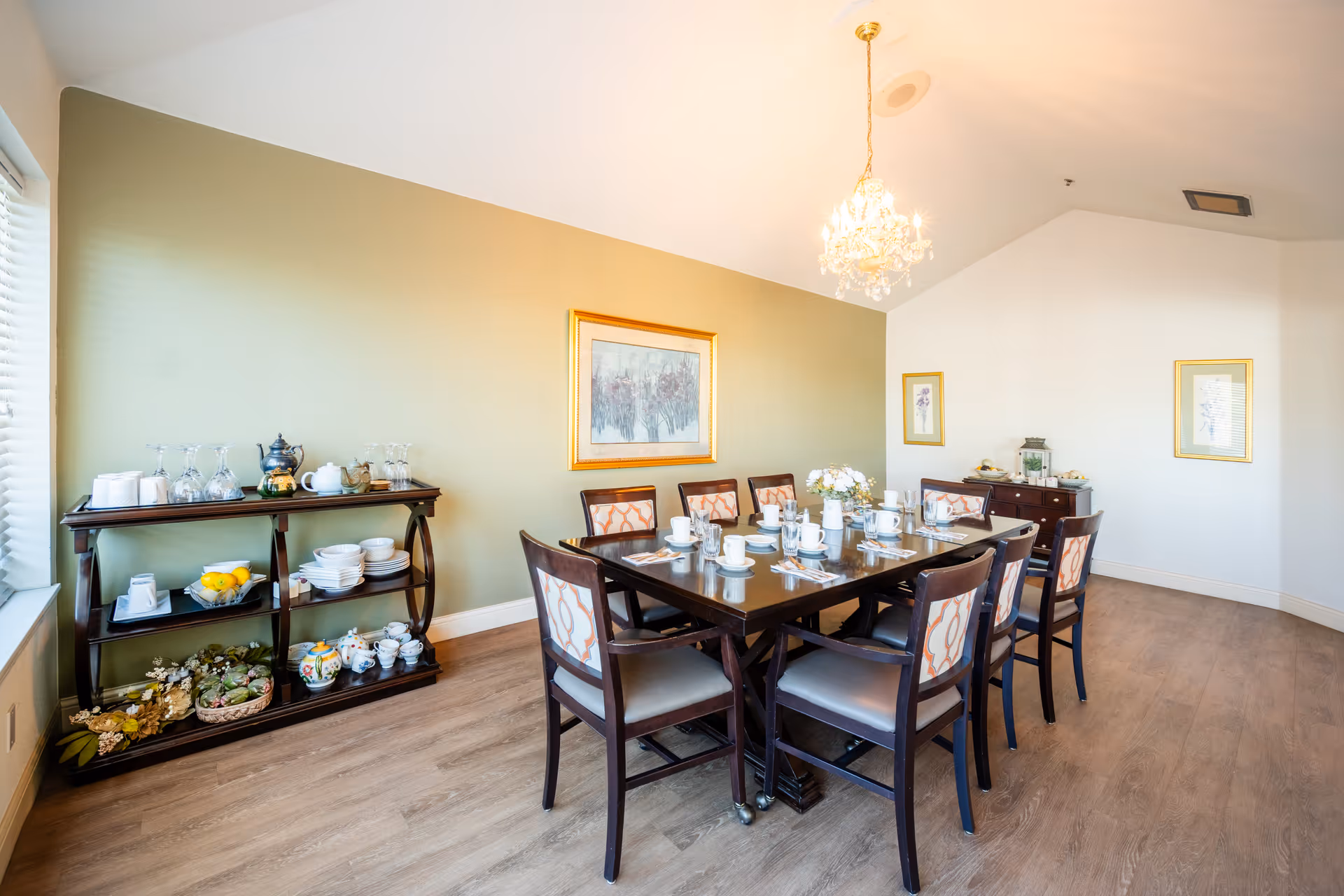 A bright dining room with a wooden table set for eight people with white cups, saucers, and silverware. The room features light wood flooring, a green accent wall with framed artwork, a chandelier hanging from the ceiling, and two dark wooden sideboards holding glassware, teapots, plates, and decorative items.