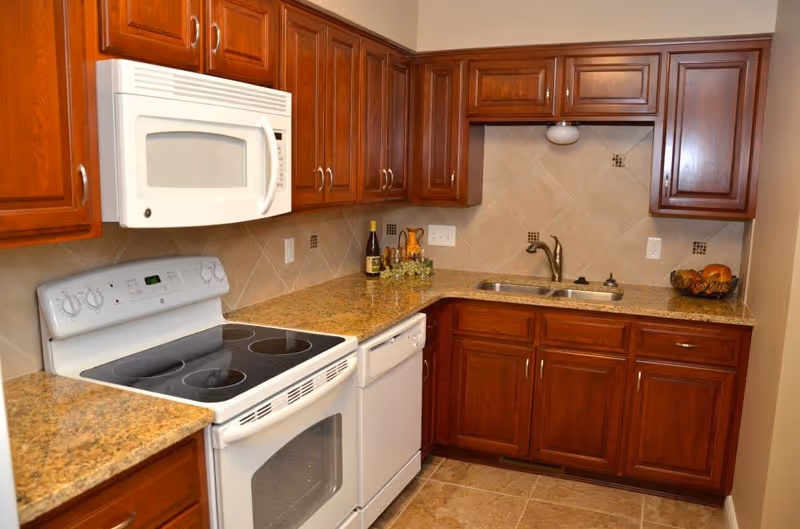 A kitchen with wooden cabinets, granite countertops, a white electric stove with an oven, a white microwave above the stove, a white dishwasher, a double sink with a faucet, and a tiled backsplash. There are some decorative items on the countertop including a bottle of wine, a small plant, and a bowl of fruit.