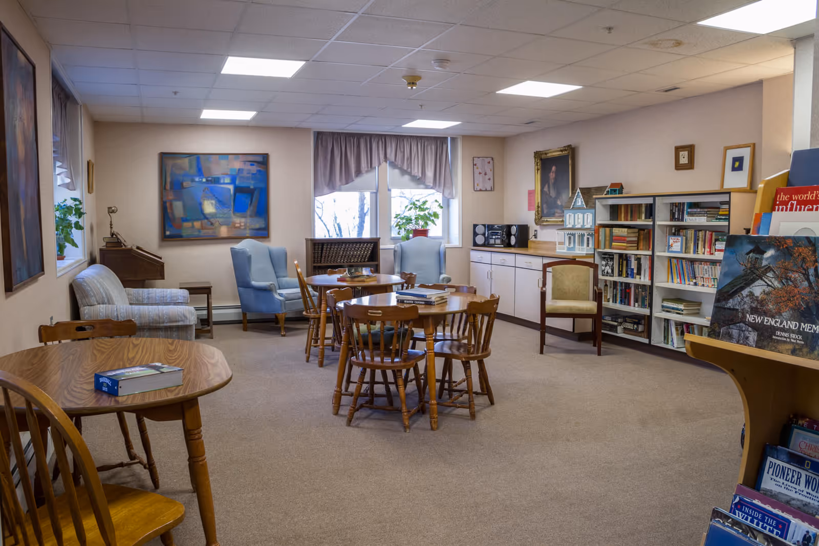 A senior living common room with round wooden tables and chairs, bookshelves, armchairs, and framed artwork.