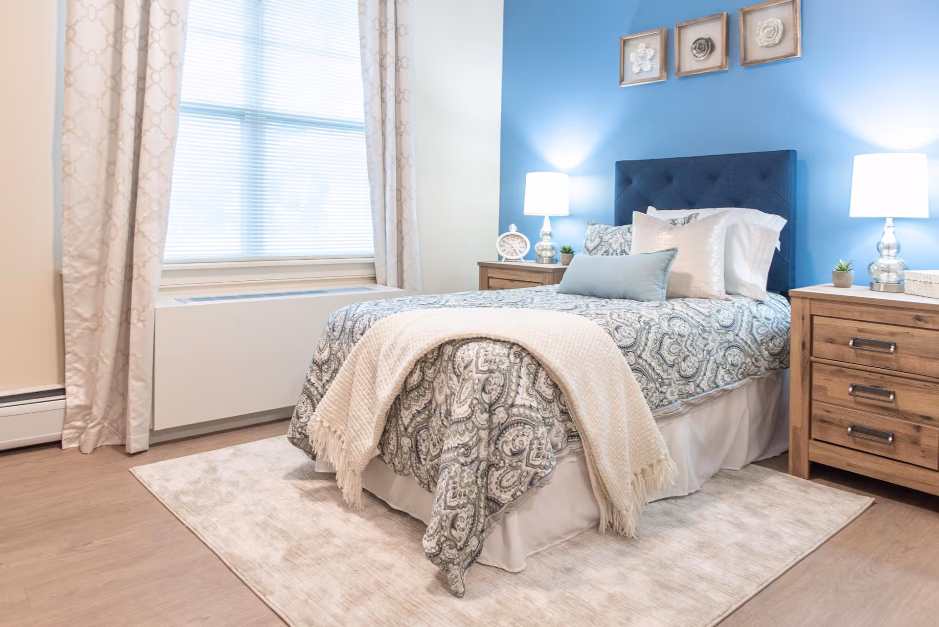 A neatly made bed in a bright bedroom with a blue accent wall, matching nightstands and lamps, and a curtained window.