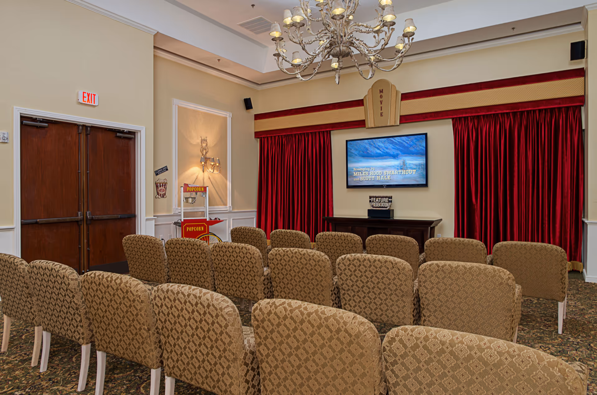 Small screening room with rows of upholstered chairs facing a TV screen flanked by red stage curtains and a popcorn cart.