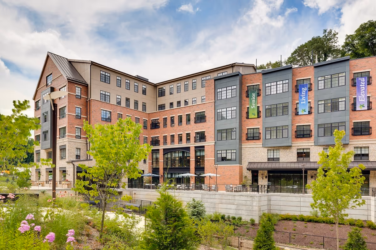Exterior view of a multi-story senior living facility building with brick and stone facade, large windows, and banners reading 'Senior', 'Living', and 'Elevated'. The foreground features landscaped greenery, trees, and a walking path under a partly cloudy sky.