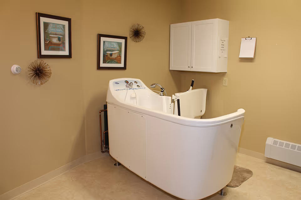 A beige-colored room with a white walk-in bathtub designed for accessibility, featuring a door and a seat inside. The room has two framed pictures of bathtubs on the wall, two decorative wall hangings, a white cabinet mounted on the wall, a clipboard, and a small heater near the floor.