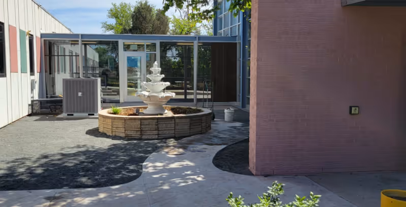 Outdoor courtyard showing a tiered fountain in a circular stone planter between building wings and a glass-enclosed walkway.