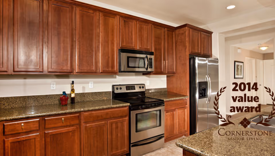 Modern kitchen with wooden cabinets, granite countertops, stainless steel appliances including a refrigerator, oven, and microwave. The kitchen is well-lit with recessed lighting and has a clean, organized appearance.